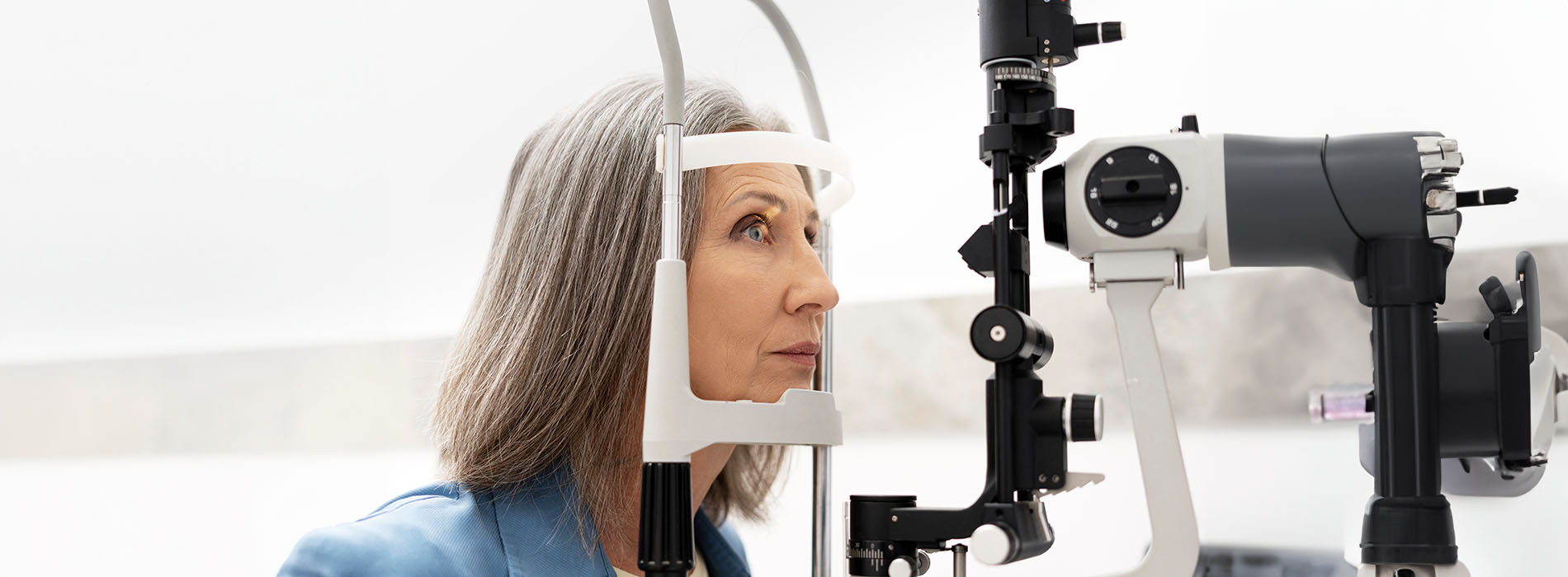 A woman with gray hair wearing a blue top stands behind an ophthalmology machine with her eyes closed, possibly during an eye examination.