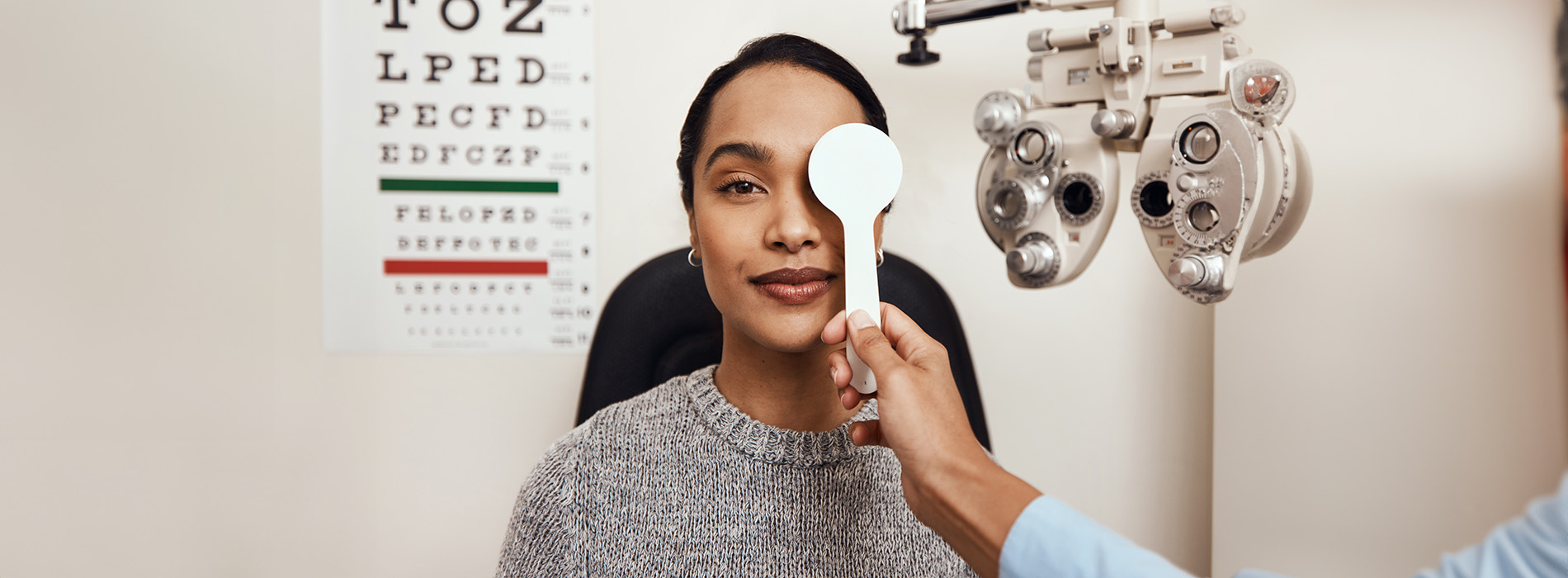 The image shows a person sitting at an eye exam machine with a large magnifying glass, while another individual appears to be assisting or performing the test by holding a pen over their eye.
