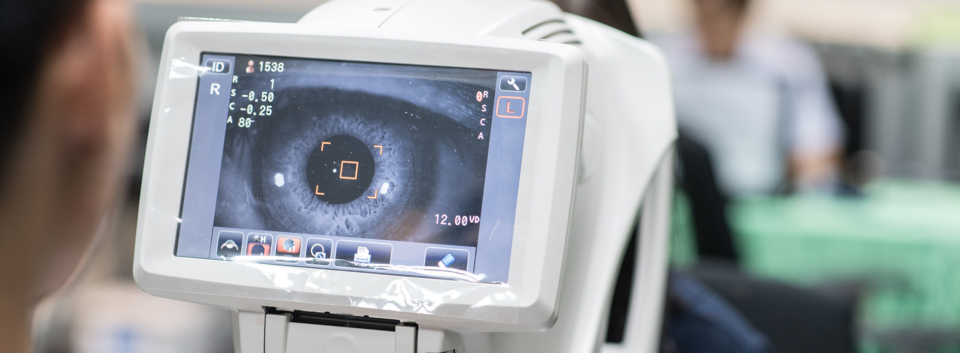The image shows a person using an eye exam machine with a display screen showing a close-up view of a human eye, likely for vision testing purposes.