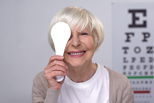 An elderly woman with white hair wearing glasses and holding a magnifying glass up to her eye.