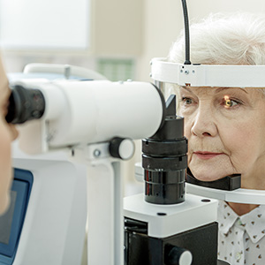 The image shows an elderly woman receiving an eye examination using a slit lamp, with her face clearly visible while she is seated at the device.