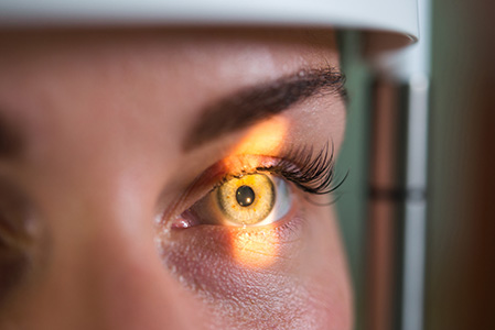 The image shows a close-up view of an eye with a bright reflection, likely from a device or light source, captured through an ophthalmic examination machine, such as a slit lamp, with a focus on the illuminated pupil and surrounding eyelashes.