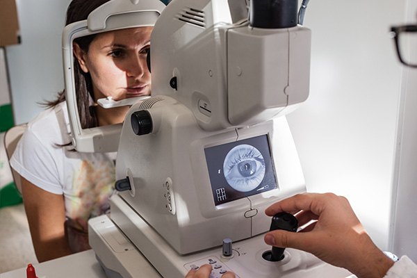 The image shows a woman sitting at an eye exam machine with a man standing beside her, adjusting the device.