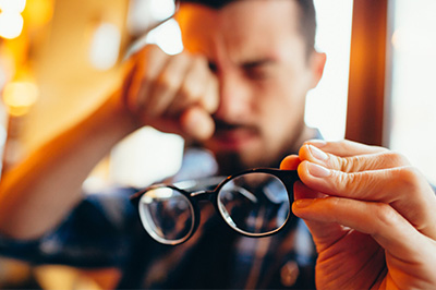 A man holding up a pair of glasses with his left hand, looking away from the camera with a concerned expression.