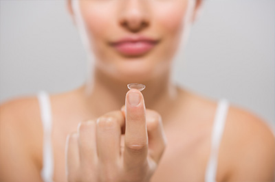 The image shows a close-up of a person s hand holding a small object with a reflective surface, which appears to be a contact lens, against their face.