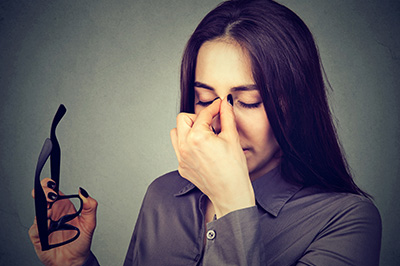 The image shows a woman holding a pair of glasses with her hand over her nose, seemingly wiping away tears or adjusting her eyewear, with a thoughtful or contemplative expression on her face.