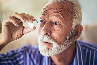 An elderly man with white hair, a beard, and mustache uses an eye dropper to put something into his eye.