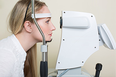 A woman with blonde hair is seated in front of an ophthalmology machine, appearing to have her eyes examined by it.