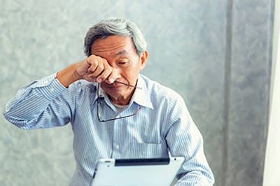 The image shows an elderly Asian man sitting at a desk with his head resting on his hand, looking downwards, seemingly tired or deep in thought, next to a laptop computer.