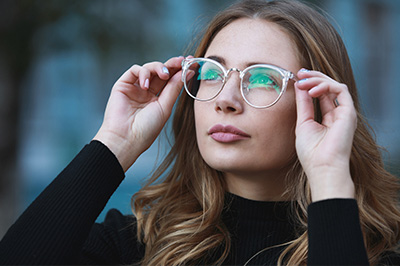 The image shows a woman wearing glasses, with her hand on her face, looking intently at something off-camera. She has light hair and is dressed in a black top.