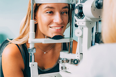A woman sitting at an eye exam machine with a device attached to her forehead, smiling and looking towards the viewer.